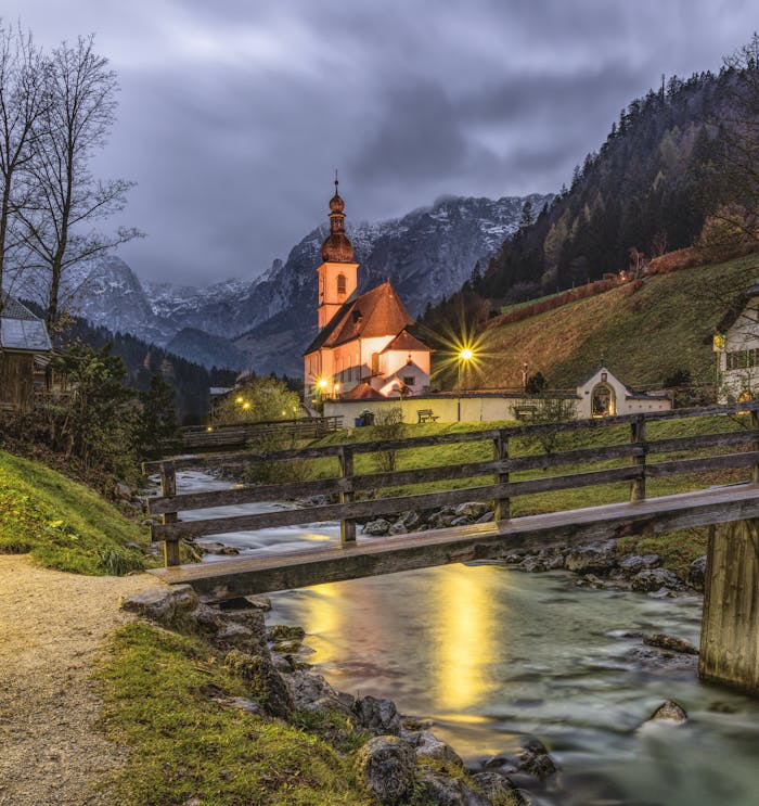 Picturesque church illuminated at twilight by a flowing river in a serene mountainous landscape.