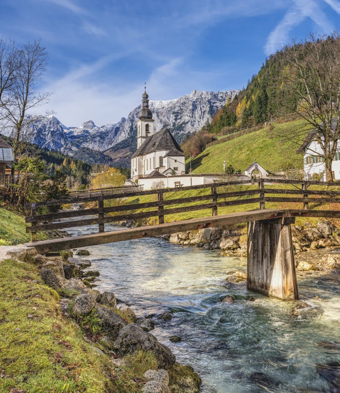 Beautiful mountain landscape with a church by a river and a wooden bridge under a clear sky.